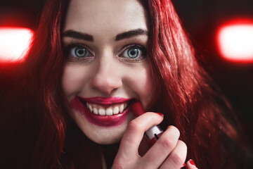 Portrait of crazy-looking teen girl with red hair she is smearing red lipstick on her face, horror concept. halloween time