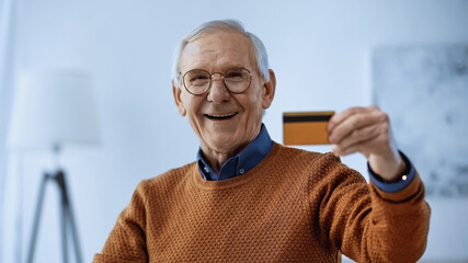 smiling elderly man in glasses holding credit card in modern living room.