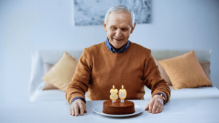 happy elderly man celebrating birthday in front of cake with burning candles in living room.