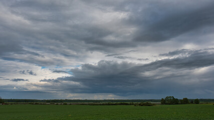 A landscape of a beautiful evening sky with storm clouds over green farmland with islands of trees and bushes, and a forest on the horizon.