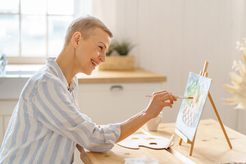 Creative hobby, digital detox. Side view of smiling mature woman painting picture at home while sitting at wooden table in kitchen, art therapy for mental health. Senior people and art concept