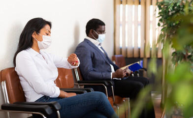 Tense latin american female entrepreneur wearing protective face mask to prevent viral infection waiting for meeting with partner, sitting in office reception, looking at her watch