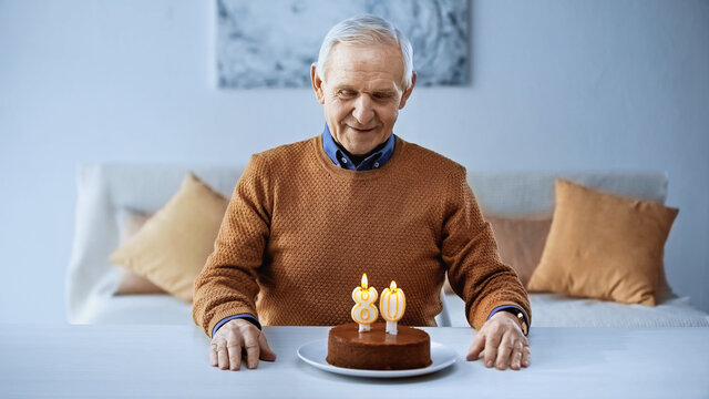 happy elderly man sitting in front of birthday cake with candles in living room.