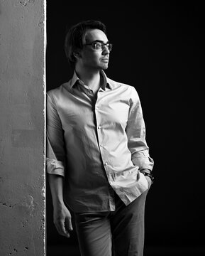 Black And White Portrait Of Adult Man In Glasses, Shirt With Rolled Up Sleeves And Jeans Standing, Leaning Against Concrete Wall With Shoulder And Looking Aside Over Black Background