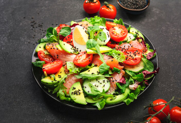 Fresh salad with salted salmon, avocado, egg, sesame seeds, olive oil, tomatoes and mixed herbs. Keto diet food. Black background, top view