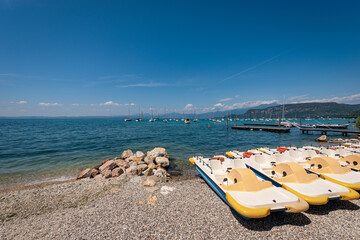 Paddle boats and sailing boats moored in front of the small Bardolino village, coast of Lake Garda (Lago di Garda). On the horizon the headland called Punta San Vigilio. Verona, Veneto, Italy, Europe.