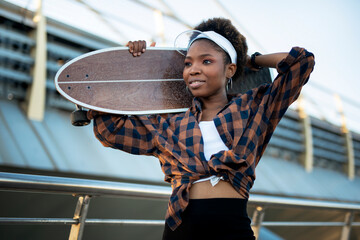 Portrait of happy african-american woman with skateboard. Young stylish woman with skateboard outdoors. © JustLife