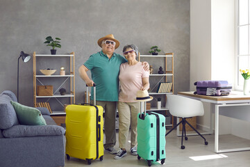 Portrait of happy senior tourists with packed suitcases standing in living room at home. Active retired elderly husband and wife ready for summer vacation. Traveling and tourism concept