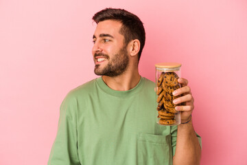 Young caucasian man holding cookies jar isolated on pink background looks aside smiling, cheerful and pleasant.