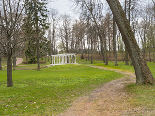 Walk in the park. A beautiful path among the trees. Green grass in early spring. In the background there is a Greek-style pillared bridge over the stream. Overcast.