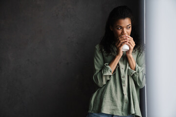 Black young woman drinking coffee while leaning on wall indoors
