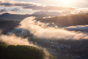Scenic aerial view of the foggy Carpathian mountains, village and blue sky with sun and clouds in morning light, summer rural landscape, outdoor travel background