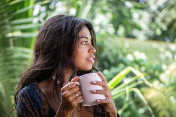 Portrait of young brunette woman freelancer drinking coffee from white cup on balcony of tropical bungalow with palm trees view