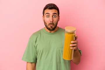 Young caucasian man holding pasta jar isolated on pink background shrugs shoulders and open eyes confused. - obrazy, fototapety, plakaty