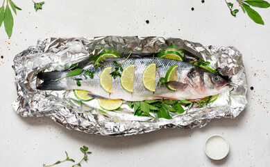 Raw fish with lemon and herbs in foil. Uncooked sea bass in foil on a gray concrete background top...