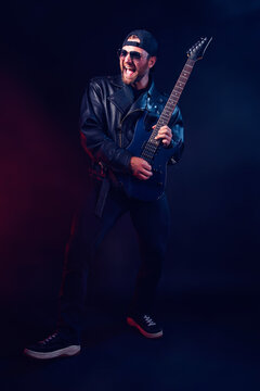Full size photo of brutal bearded Heavy metal musician in leather jacket and sunglasses is playing electrical guitar very emotionally. Shot in a studio on dark background