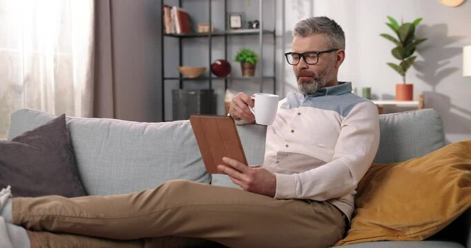 Middle-aged handsome mature Caucasian man in glasses resting on sofa in good mood drinking tea or coffee watching videos on tablet device on internet online in living room, leisure, home concept