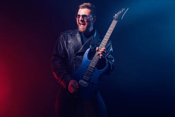 Obraz na płótnie Canvas Brutal bearded Heavy metal musician in leather jacket and sunglasses is playing electrical guitar. Shot in a studio on dark background with smoke