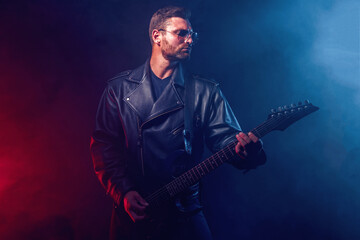 Obraz premium Brutal bearded Heavy metal musician in leather jacket, cap and sunglasses is playing electrical guitar. Shot in a studio on dark background