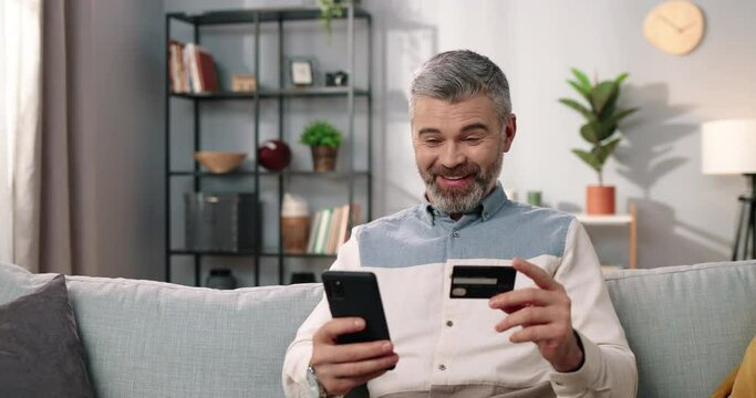 Close up of joyful Caucasian middle-aged bearded man sitting on couch in cozy room tapping on smartphone using credit card buying online in good mood satisfied making YES gesture, e-commerce, shopping