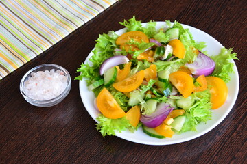 vegan salad with yellow tomatoes, red onion, cucumber and green salad leaves with pink salt on the wooden kitchen table