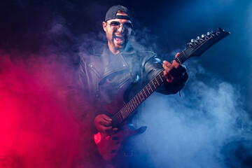 Brutal bearded Heavy metal musician in leather jacket and sunglasses is playing electrical guitar. Shot in a studio on dark background with smoke