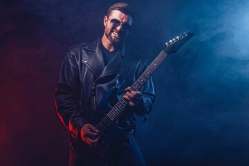 Obraz premium Brutal bearded Heavy metal musician in leather jacket and sunglasses is playing electrical guitar. Shot in a studio on dark background with smoke