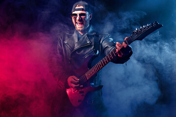Brutal bearded Heavy metal musician in leather jacket and sunglasses is playing electrical guitar. Shot in a studio on dark background with smoke