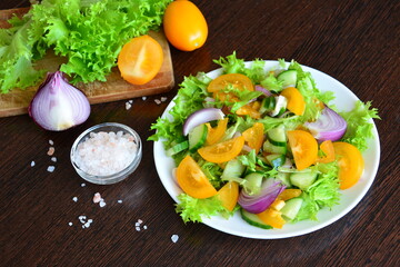 vegan salad with yellow tomatoes, red onion, cucumber and green salad leaves with pink salt and cutting board with fresh vegetables on the wooden kitchen table