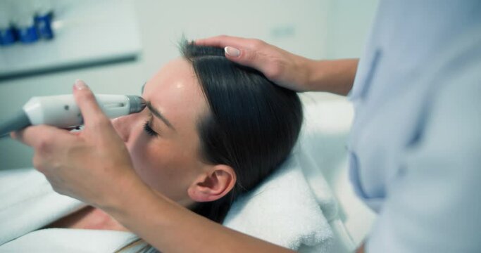 Close-up a female client in a bathrobe is laying in the cosmetology bed while having a non-injection bio-revitalization procedure on her upper eyelid, brow, temple by using the Dermadrop TDA device