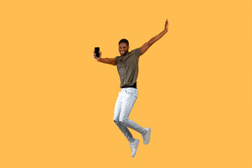 Fototapeta na wymiar Joyful african americna man jumping over yellow background and showing smartphone with black screen to camera.
