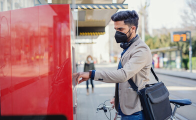 Young business man commuter buying ticket on the way to work outdoors in city, coronavirus concept.