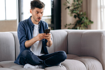 Obraz premium Shocked confused young caucasian man, in casual wear, sitting on sofa in lotus position, uses smart phone, receiving an unexpected message, texting with friends, looking at screen in surprise