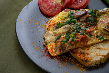 Rustic omelette made with mushrooms and spring onions, sprinkled with smoked paprika and curcuma, on a slice of grilled bread. Raw mushrooms and slices of tomato, on a black ceramic plate.