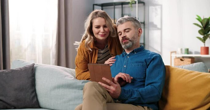 Portrait of handsome Caucasian middle-aged bearded grey-haired man sitting on sofa in room surfing internet on tablet browsing and choosing something with his beautiful happy wife, family concept