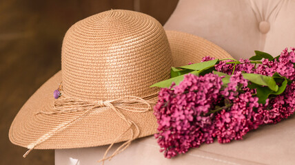 Decorative composition with a bouquet of lilacs and a beige wicker hat. Beach wicker hat, flowers, close-up. Lilac bouquet. Cozy atmosphere of relaxation and romance. Summer background. 