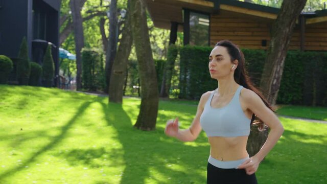 Slow motion woman with ponytail wearing sports top and wireless earphones jogging in empty park with street cafes on background. Tracking shot female athlete training outside in summer