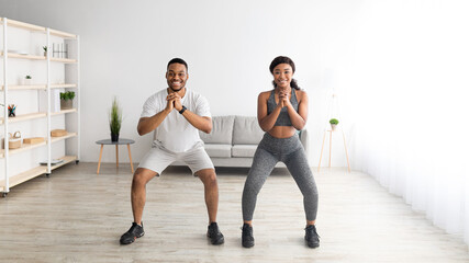 Domestic strength workout. Motivated black lady and her male partner doing squats together at home, banner design