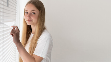 A portrait of a blonde girl standing by the window looks behind the blinds to the left of the space for the text. White background in the office. White wall and woman on the background.