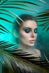 Beauty woman in palm leaves wet makeup, tropical portrait girl in green swimsuit in branches palm tree in studio, smoke and raindrops on glass. Sexy woman with bright green makeup
