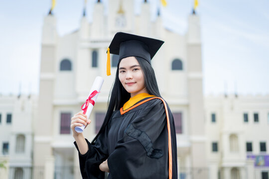 A young beautiful Asian woman university graduate in graduation gown and mortarboard holds a degree certificate stands in front of the university building after participating in college commencement