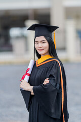 A young beautiful Asian woman university graduate in graduation gown and mortarboard holds a degree...