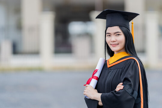 A young beautiful Asian woman university graduate in graduation gown and mortarboard holds a degree certificate stands in front of the university building after participating in college commencement