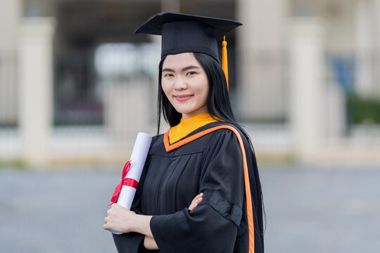 A young beautiful Asian woman university graduate in graduation gown and mortarboard holds a degree certificate stands in front of the university building after participating in college commencement