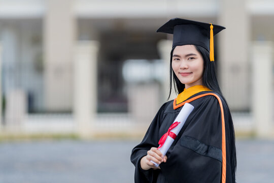 A young beautiful Asian woman university graduate in graduation gown and mortarboard holds a degree certificate stands in front of the university building after participating in college commencement
