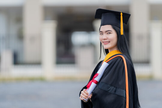 A young beautiful Asian woman university graduate in graduation gown and mortarboard holds a degree certificate stands in front of the university building after participating in college commencement