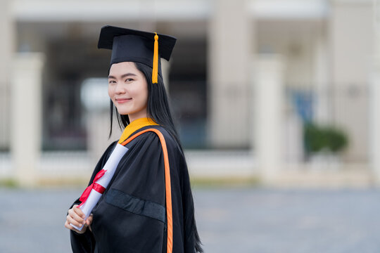 A young beautiful Asian woman university graduate in graduation gown and mortarboard holds a degree certificate stands in front of the university building after participating in college commencement