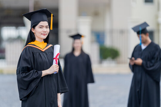 A young beautiful Asian woman university graduate in graduation gown and mortarboard holds a degree certificate stands in front of the university building after participating in college commencement
