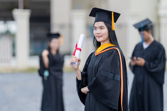 A young beautiful Asian woman university graduate in graduation gown and mortarboard holds a degree certificate stands in front of the university building after participating in college commencement