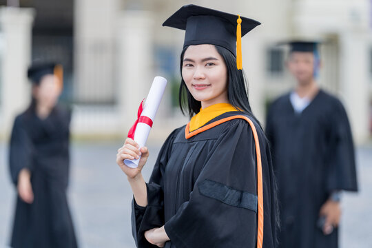 A young beautiful Asian woman university graduate in graduation gown and mortarboard holds a degree certificate stands in front of the university building after participating in college commencement